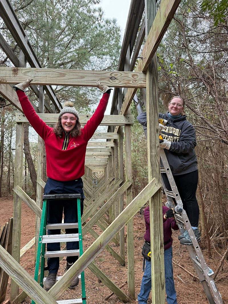 Two students pose standing on ladders next to wooden frame structure with power tools in hand. 
