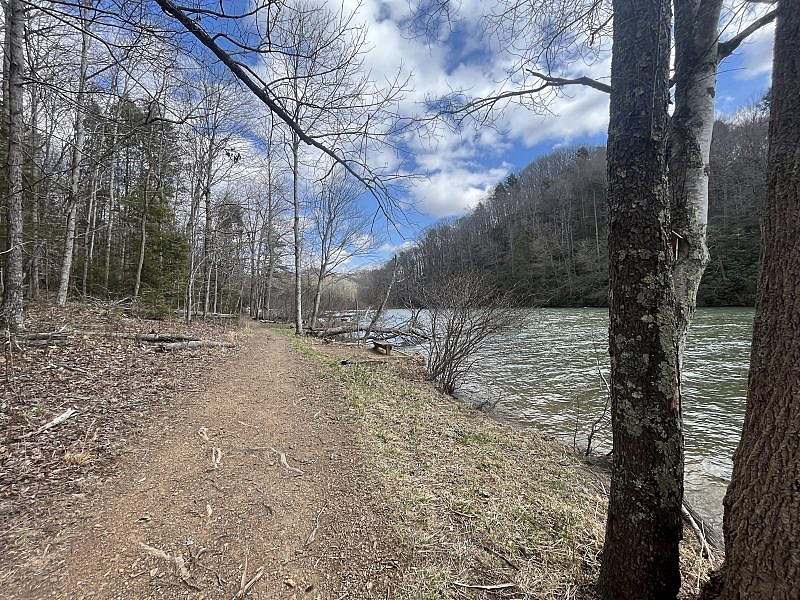A dirt trail runs alongside a calm river, bordered by leafless trees under a partly cloudy sky.