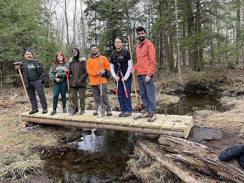6 people stand with trail tools on a  newly built bridge that spans a stream, providing more accessible trail access
