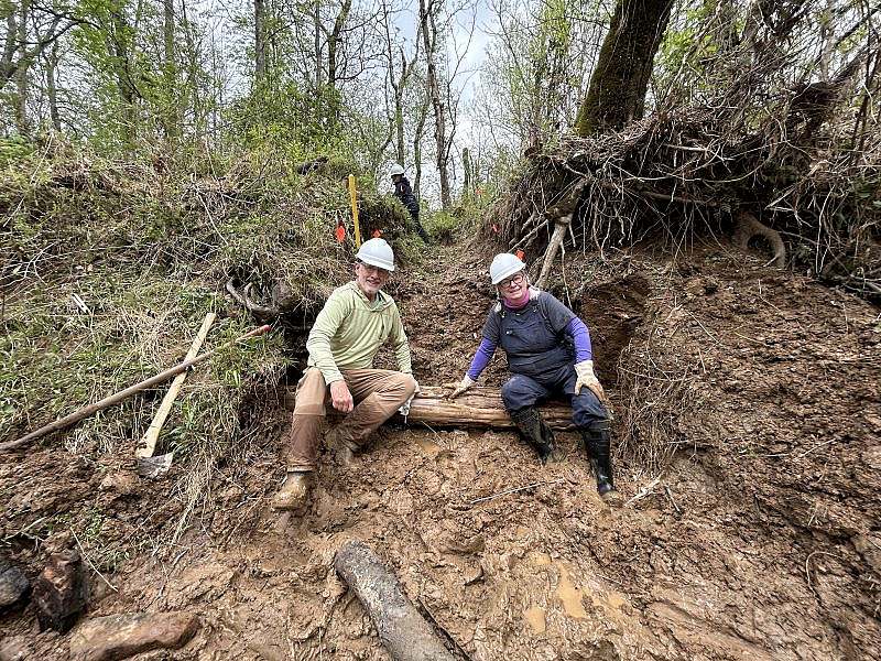 Two smiling volunteers, ankle-deep in mud and dressed in work clothes and muck boots, sit on a newly reinforced trail section designed to prevent erosion.