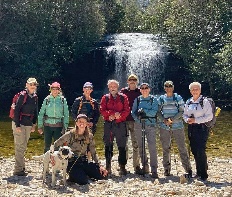 Nine hikers and a dog stand in front of a waterfall on the Glen Falls Trail in Nantahala National Forest. Surrounded by lush greenery, the group poses in outdoor gear with backpacks and trekking poles, smiling on a sunny day.