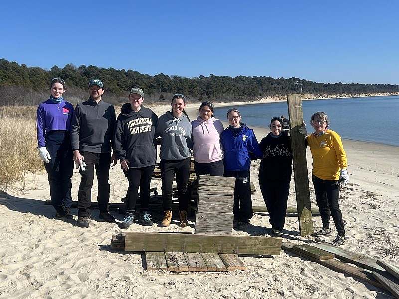 8 people stand on beach posing with old wood from a bridge that they replaced.
