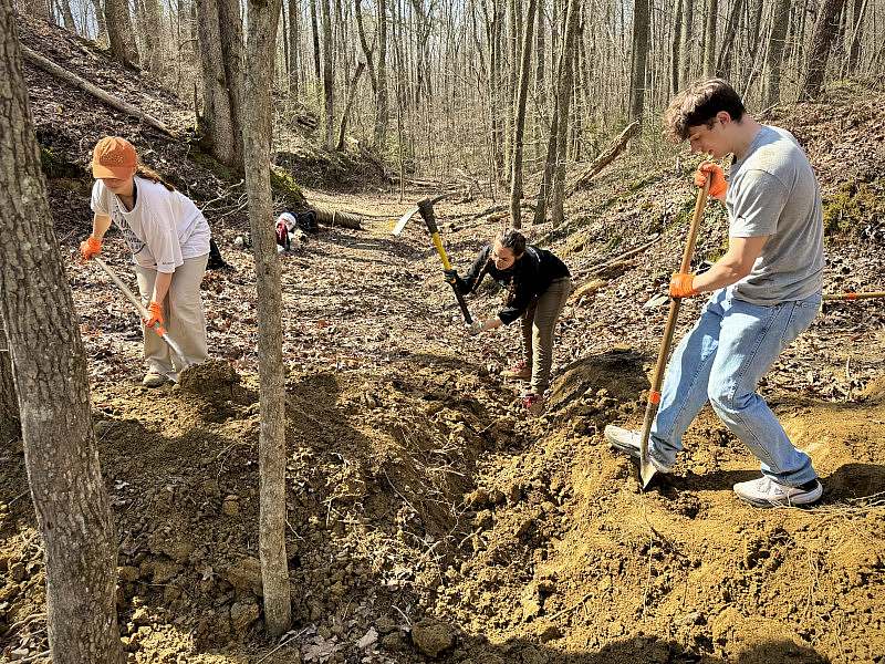 3 students use shovels and a pickaxe to break apart dirt. 
